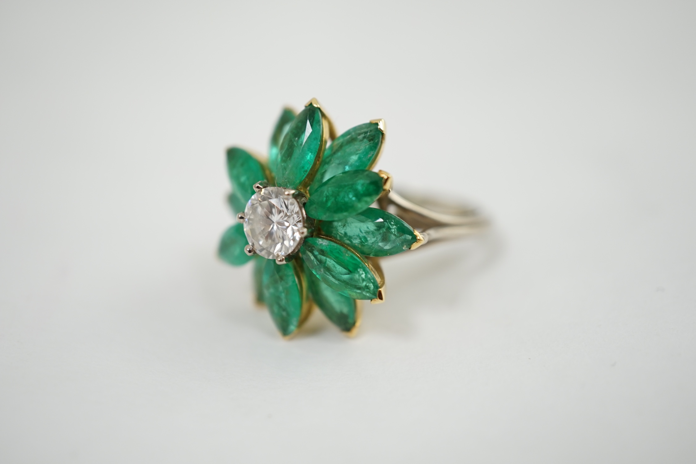 An 18ct white gold and yellow gold, emerald and diamond flowerhead dress ring, the central circular cut diamond surrounded by eleven navette shaped emerald 'petals', all claw set